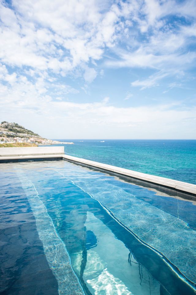 Depuis le 3e étage, la piscine semble fusionner avec la mer. Une ouverture sur la pièce de vie permet de laisser les reflets de l'eau se propager entre les espaces.