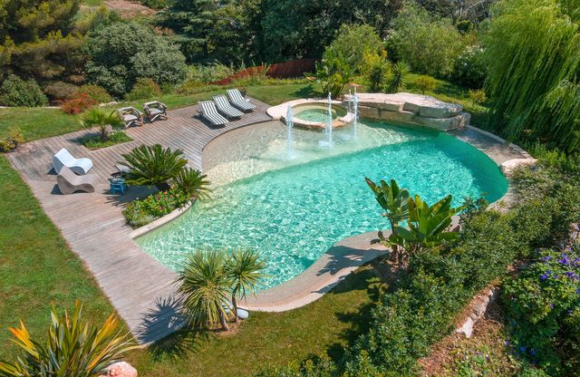 Piscine construite en béton armé monobloc de forme libre, d'une superficie de 80 m2 dont une plage immergée de 20 m2. Le spa de 2m attenant à la piscine s'y déverse. Deux revêtements minéraux : Marbreroc® Sable avec option Impérial, à base de purs cristaux de marbre de carrare, pour l'ensemble de la piscine et Naturalroc® Sable, à base de micro gravillons de rivière, pour les bords naturels. Piscine connectée et autonome avec l'Hydrocapt Pilot et le système de nettoyage automatique intégré : Diffaclean. Cette piscine est caractérisée aussi par sa cascade, trois geysers et des jeux de lumières. Diffazur Piscines.