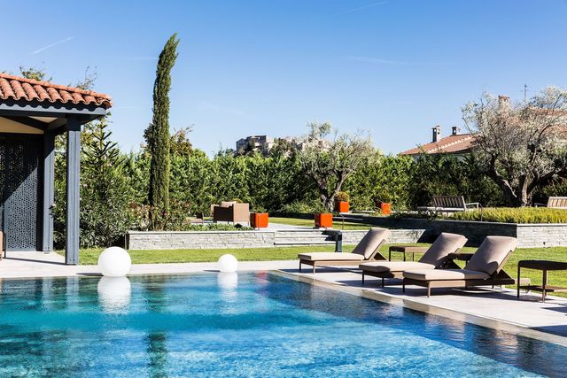 Au premier plan, la terrasse de piscine en pierre naturelle, est construite en L, pour permettre l'accès facile sur la largeur du bassin et l'aménagement d'un espace détente sur la longueur. En arrière-plan, une petite terrasse cosy en pierre est confortablement meublée, pour se détendre au calme. Réalisation Nature & Prestige.